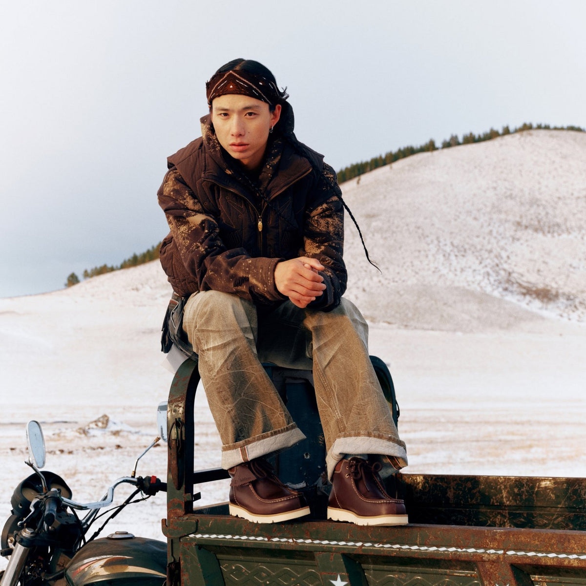 Clarks Walla Orkhon in British Tan, lifestyle shot of person in vest and cuffed jeans sitting on a cart in snowy hills