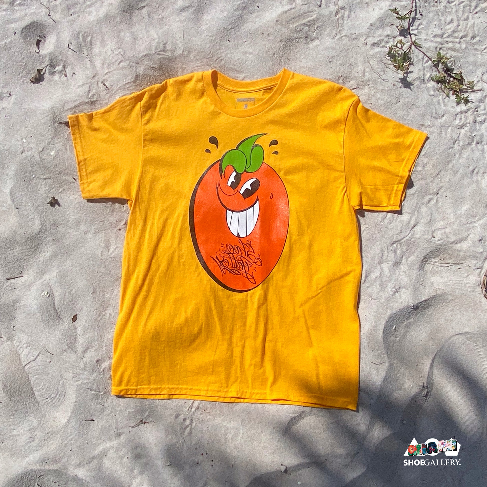 An orange t-shirt with a graphic print of a smiling orange character on the front, laid out on a sandy surface.