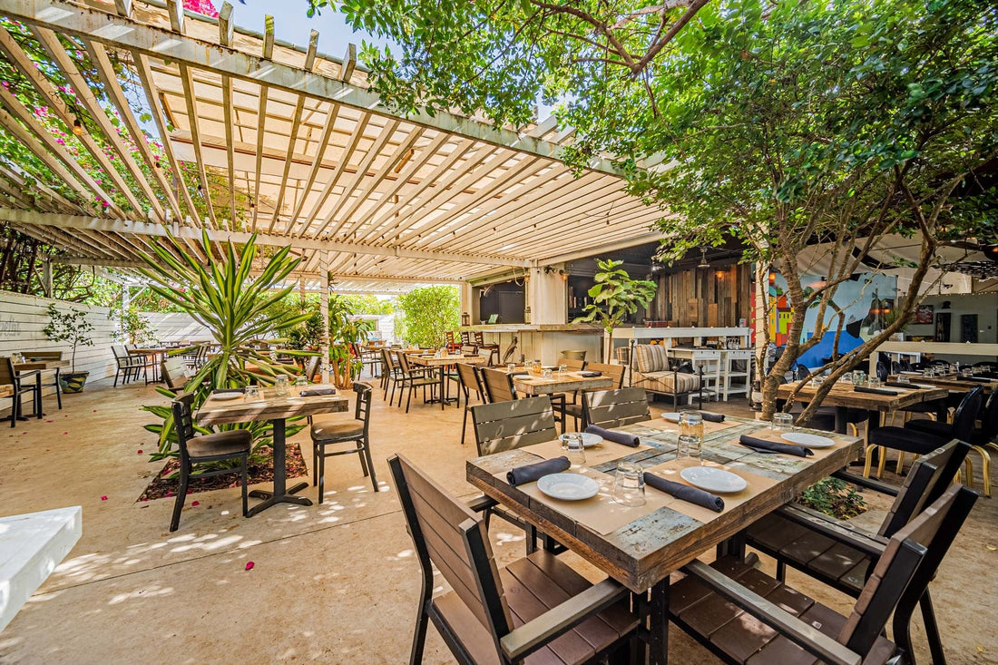 Shaded patio with wooden tables set with plates, napkins and glasses under a white pergola, surrounded by trees and tropical plants