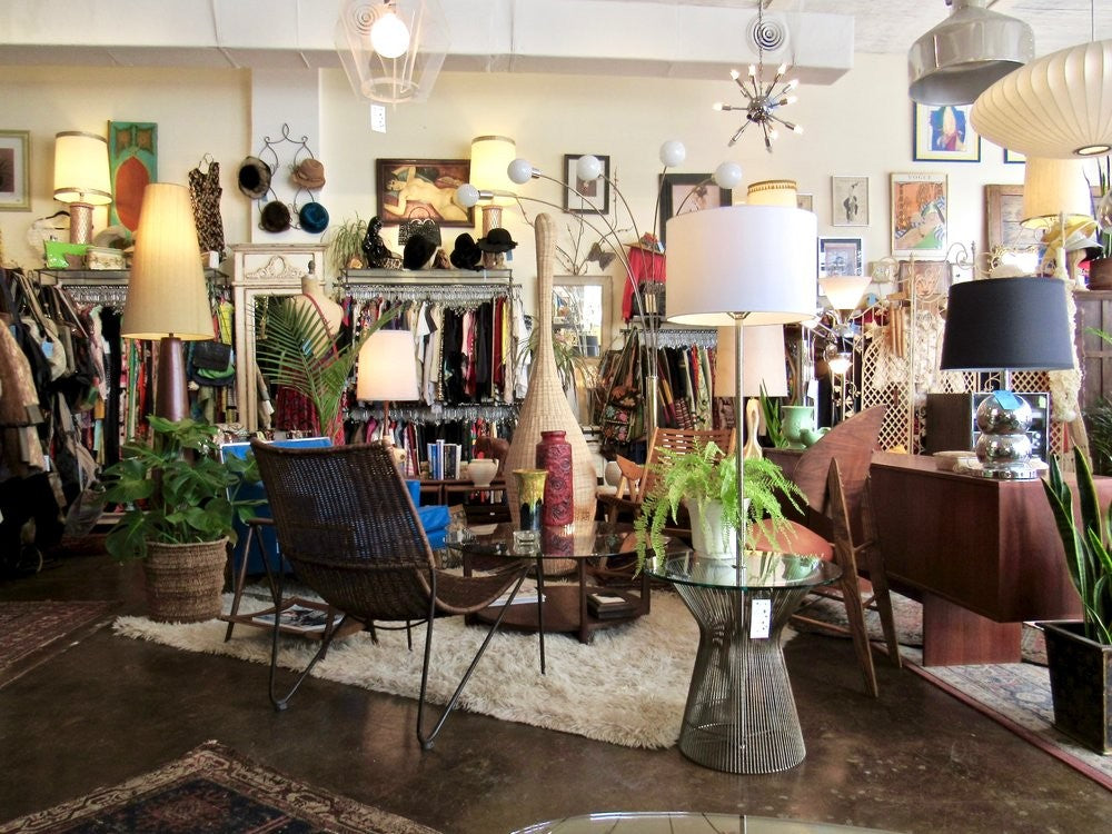 Fly Boutique interior with midcentury lamps, a wicker lounge chair, glass tables and racks of vintage clothing and hats