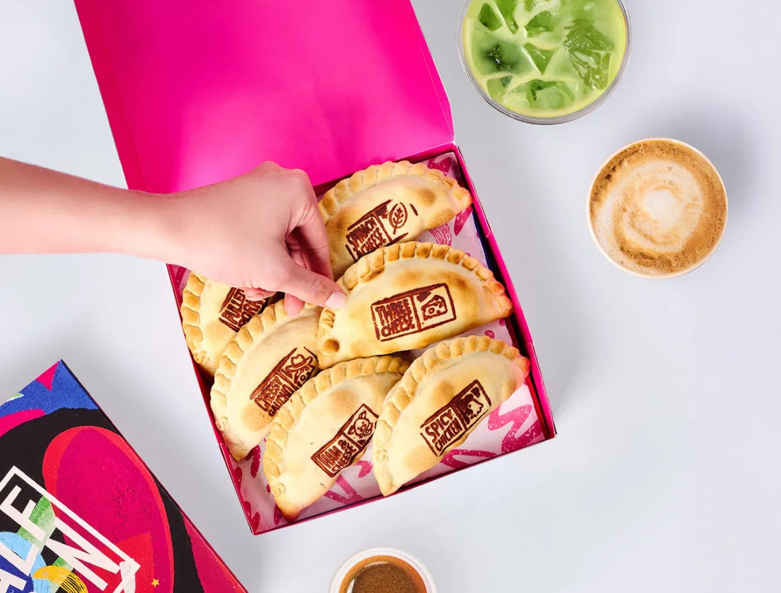 A hand picks a baked empanada from a pink box of flavor-stamped Half Moon empanadas, next to an iced green drink and a latte