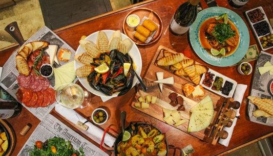 Tapas spread on a wooden table: mussels with toast, charcuterie, cheese boards, croquetas, olives, a fish dish and white wine