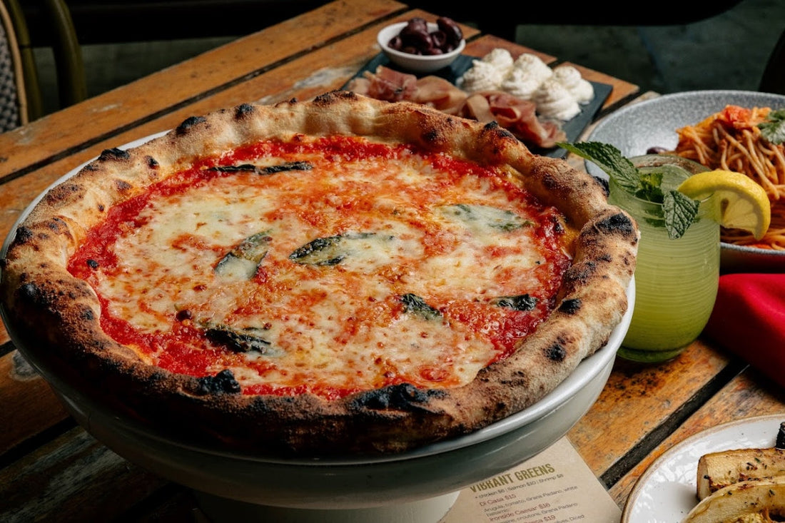 Margherita pizza with a blistered wood-fired crust, tomato, melted mozzarella and basil on a wooden table with drinks and pasta
