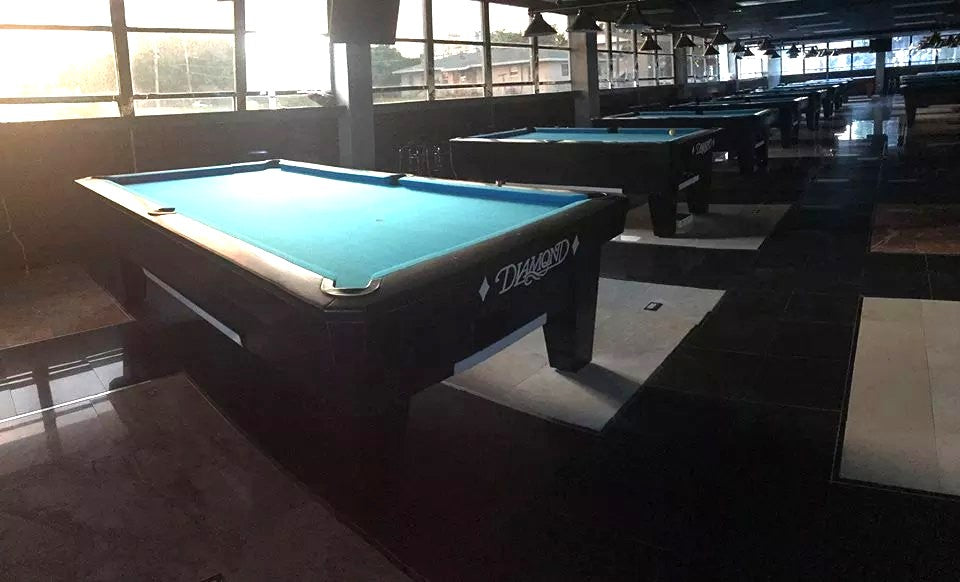 Rows of blue felt Diamond pool tables stretching back through K&K Billiards, with daylight from the windows along the far wall