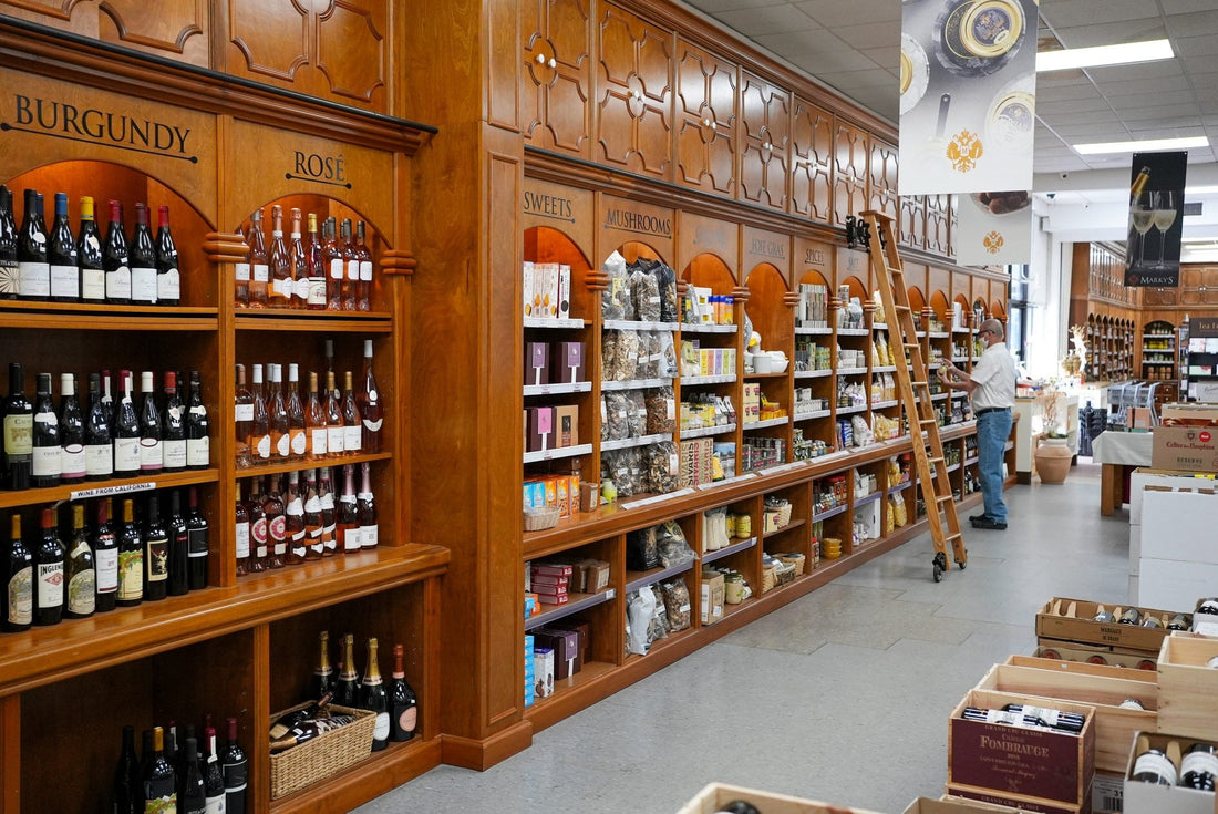 Aisle inside Marky's Gourmet with wood shelving labeled Burgundy, Rosé, Sweets and Mushrooms, and wine cases on the floor
