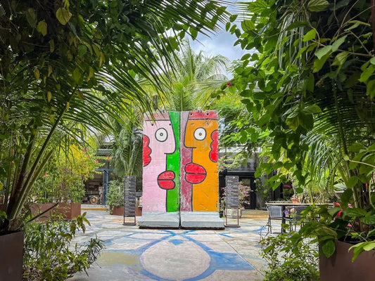 Two Berlin Wall segments painted with bold cartoon faces by Thierry Noir stand in a palm-shaded plaza at Miami Ironside.