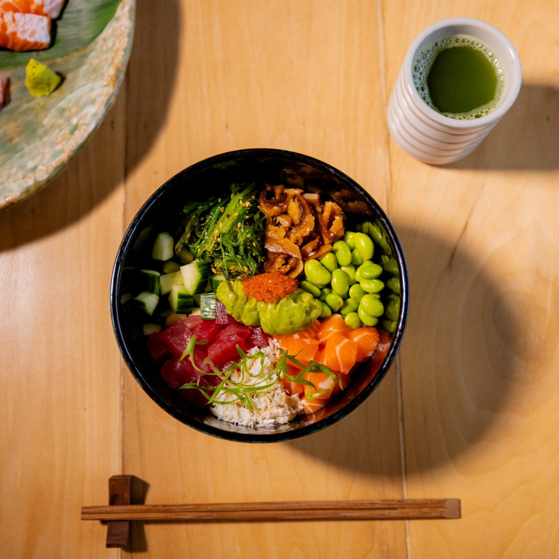 Donburi bowl of tuna, salmon, crab, tobiko, shiitake, edamame, cucumber, seaweed salad and avocado next to a cup of green tea