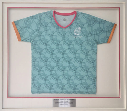 Teal North Bay Village Soccer jersey mounted in a white shadow box frame with a pink inner border and engraved plaque