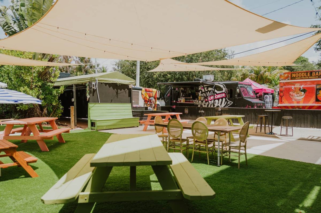 Open-air courtyard at Upper Eastside Hangout with green turf, picnic tables, cream shade sails and food trucks along the back