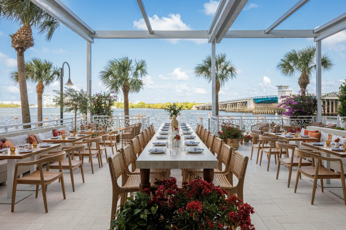 YAYA's waterfront terrace under an open white pergola, with a long communal table, woven wood chairs, palm trees and a bridge over the bay