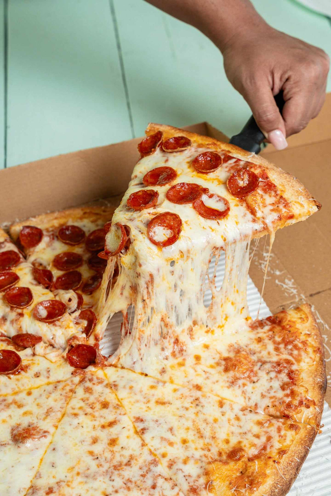 A hand lifts a pepperoni slice from a half pepperoni, half cheese pizza in its box, melted mozzarella stretching below