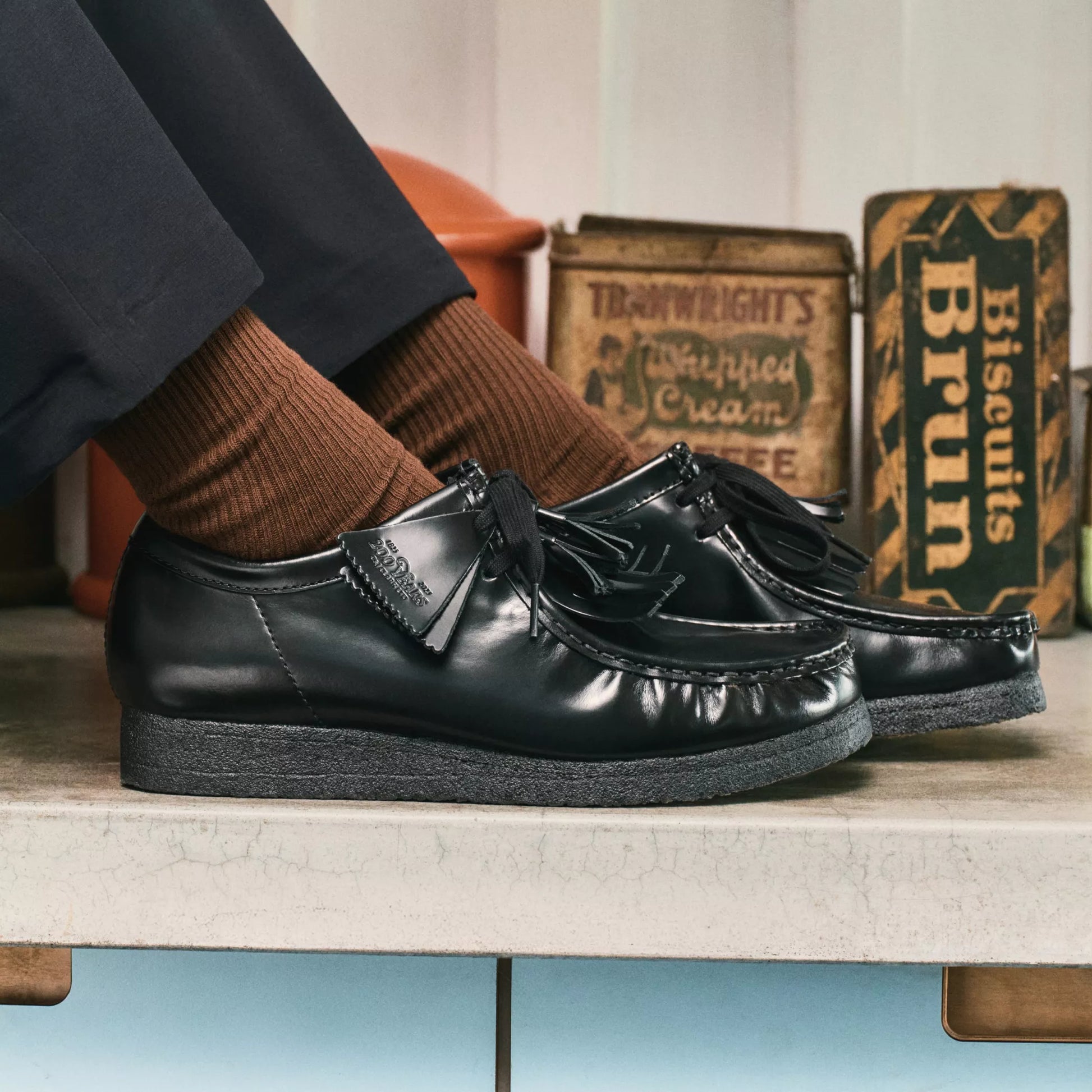Black loafers with tassels worn by a person, with vintage-style boxes in the background.