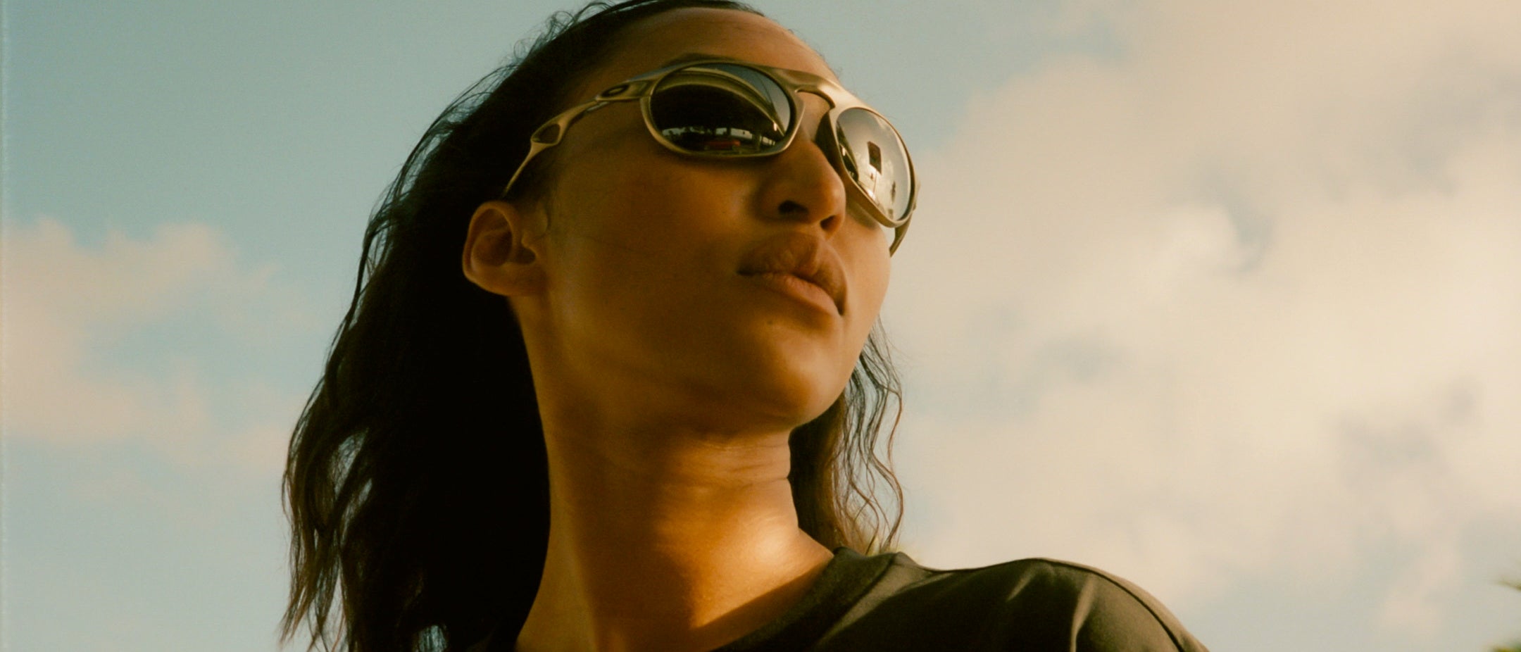 A close-up shot of a woman wearing round mirrored sunglasses against a cloudy sky background.